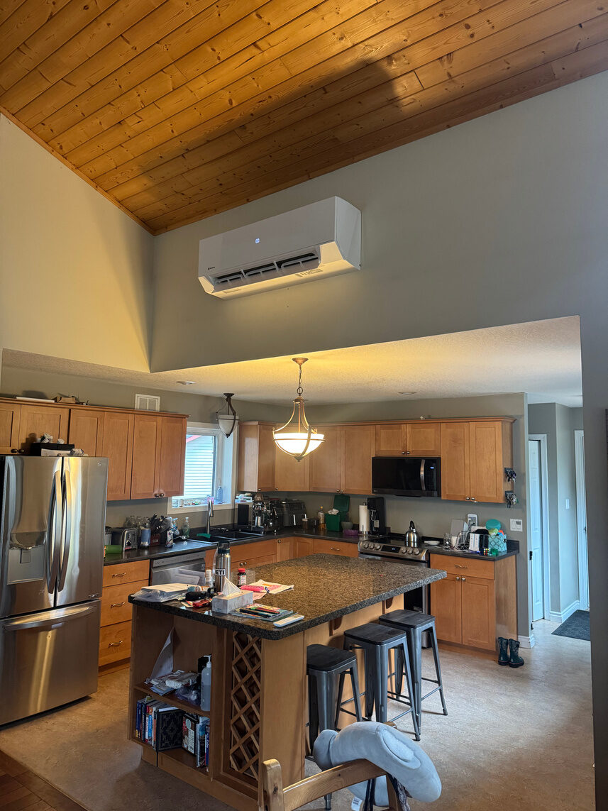 Ductless heat pump head mounted high on a wall above an open kitchen with a vaulted wood ceiling