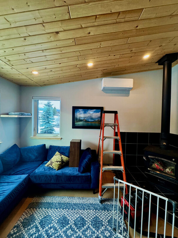 Ductless heat pump head mounted on a living room wall near a wood stove, with a stepladder below it