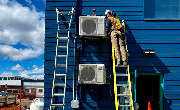 Technician on a ladder working on two wall-mounted heat pump outdoor units on a blue commercial building