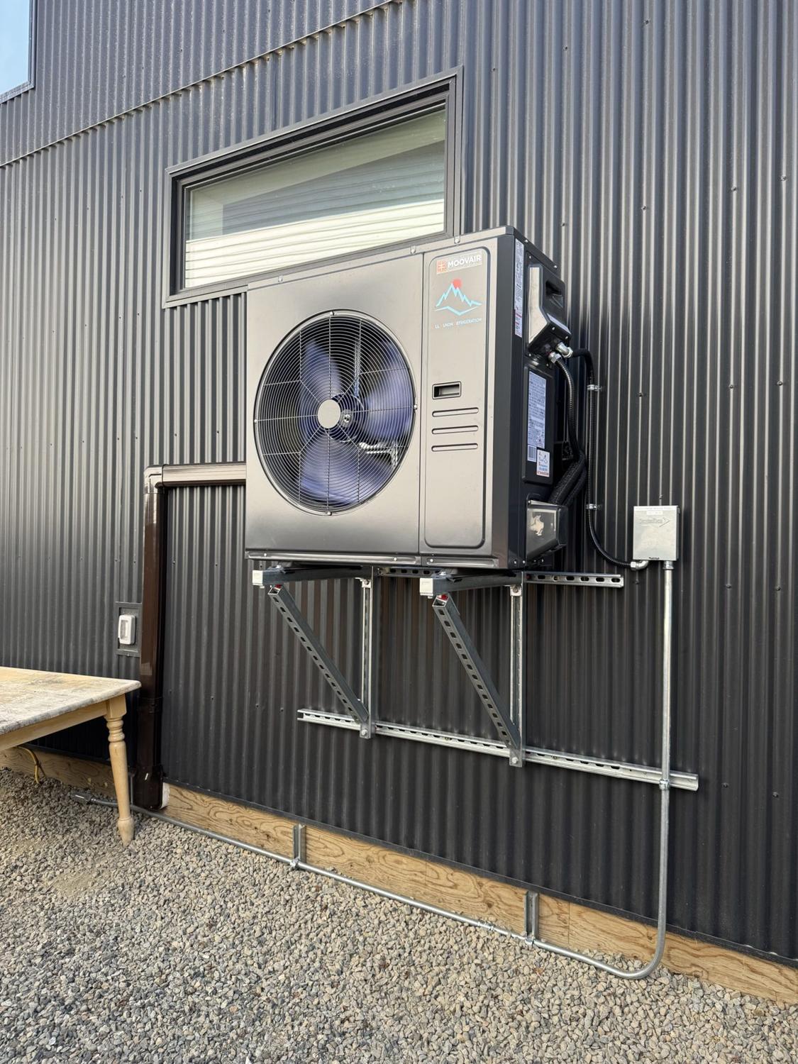 Cream heat pump outdoor unit mounted on galvanized steel wall brackets against dark corrugated metal siding, with a window above, an electrical box to the right and conduit running down to a gravel strip over timber edging