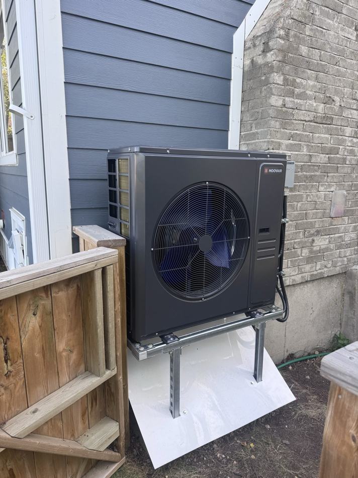 Dark MOOVAIR heat pump outdoor unit raised on a tall steel stand over a white pad, tucked between blue lap siding and a grey brick chimney with a wooden fence in the foreground