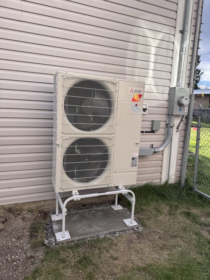 Cream Mitsubishi Electric two-fan heat pump outdoor unit on a low white steel stand over a concrete pad, beside beige lap siding with an electrical disconnect and hydro meter on the wall