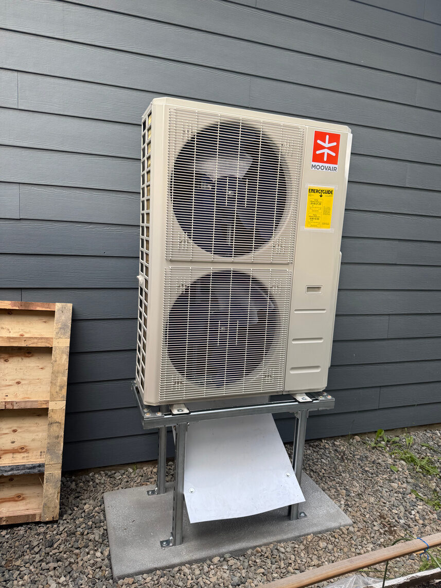 Large two-fan heat pump outdoor unit mounted on a steel stand over gravel beside dark grey siding