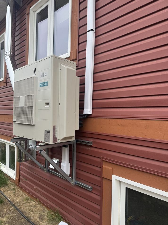 Cream Fujitsu Airstage heat pump outdoor unit on steel wall brackets against deep red lap siding, with a white line-set cover running down the wall to a window