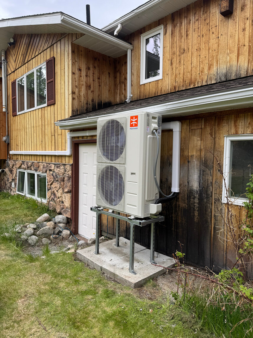 Heat pump outdoor unit on a tall steel stand at the side of a wood-sided house