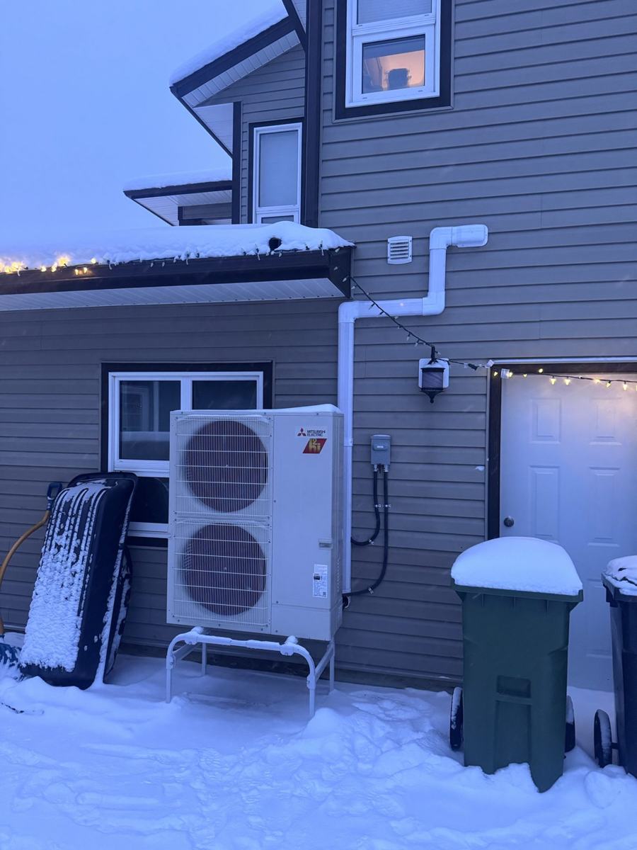Mitsubishi heat pump outdoor unit on a white stand beside a snow-covered house at dusk, with warm lights in the windows and string lights along the roof edge