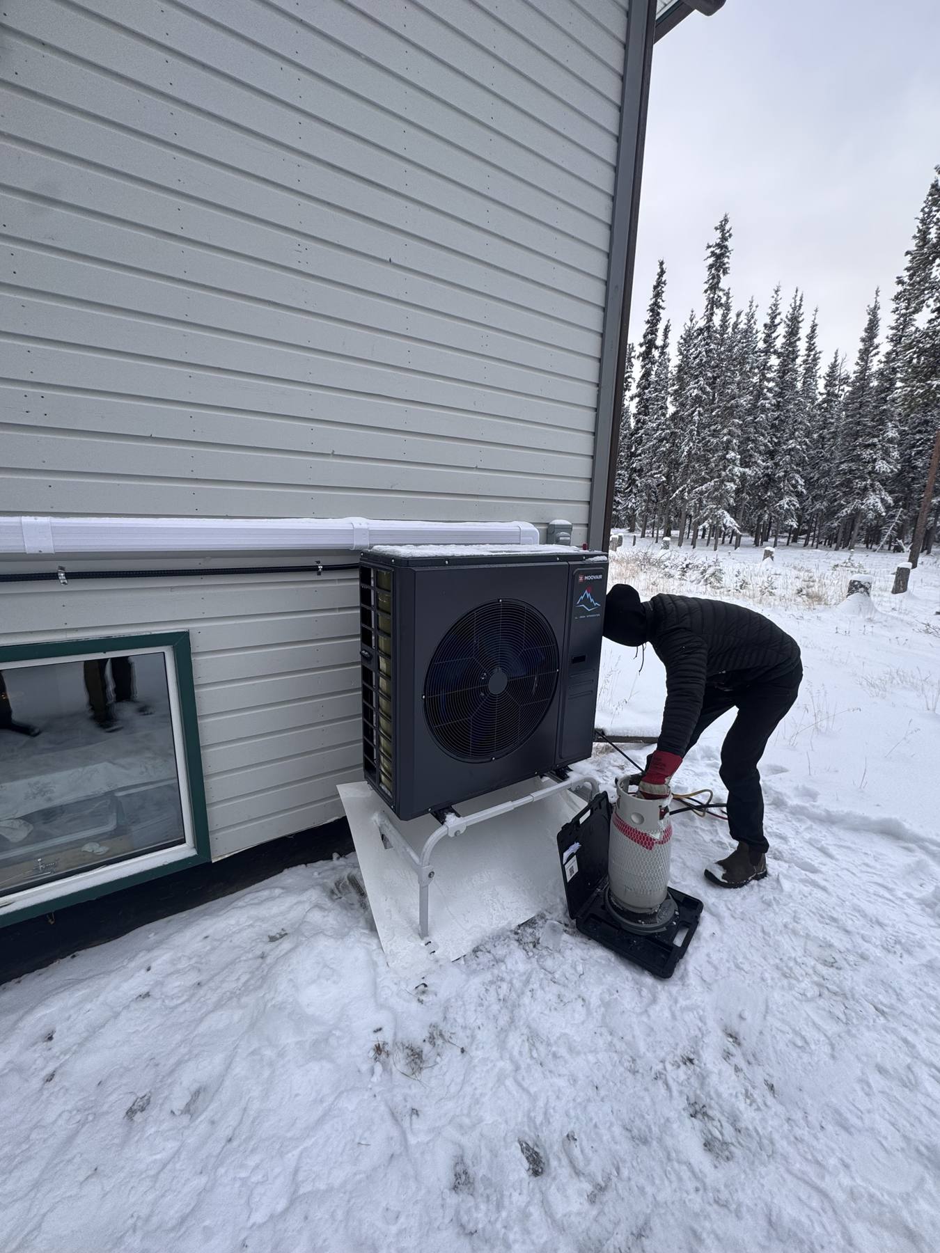 Technician commissioning a heat pump outdoor unit in deep snow beside a building, with spruce forest behind